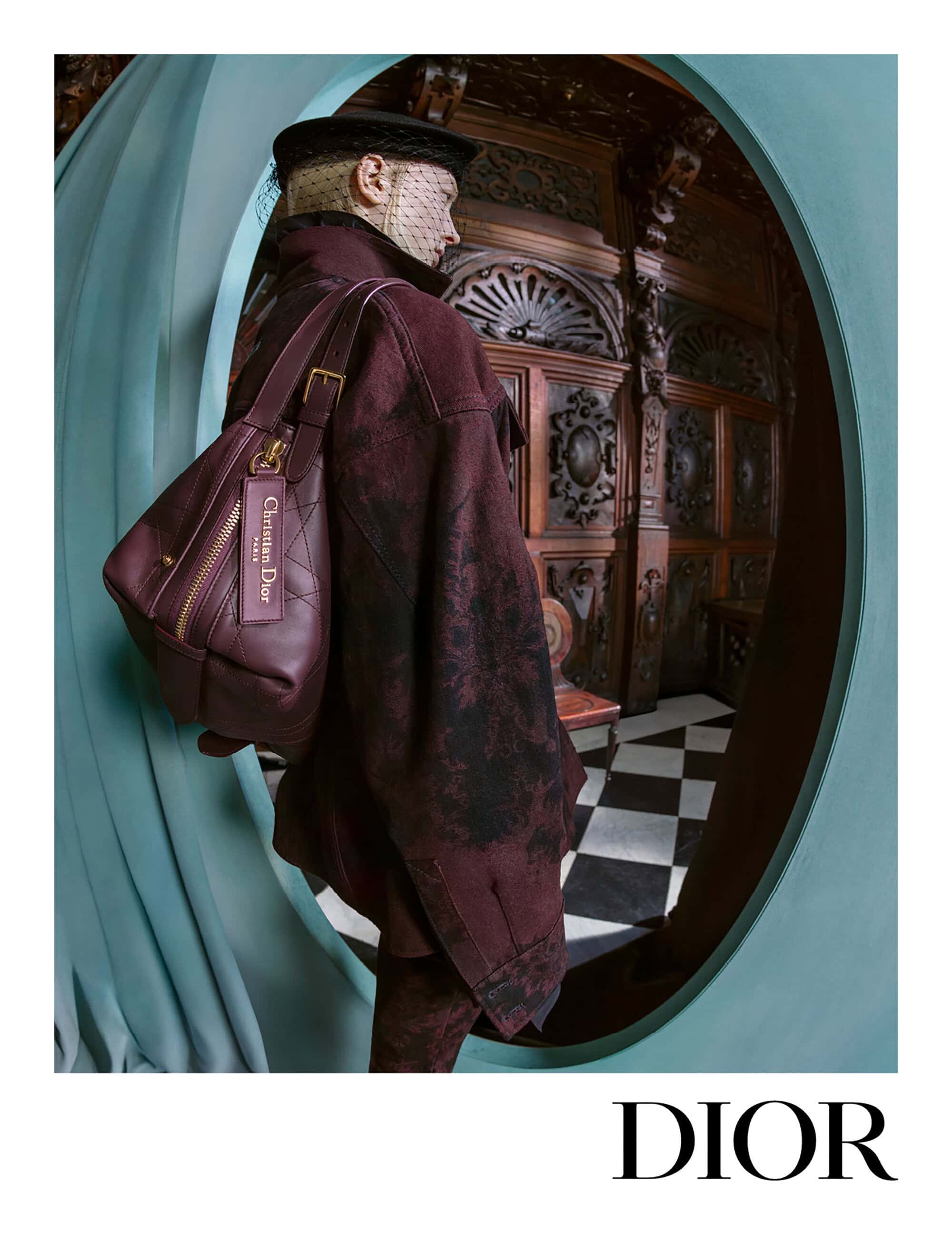 Model wearing a textured burgundy jacket and matching handbag, framed within an ornate wooden interior through a curved teal archway.