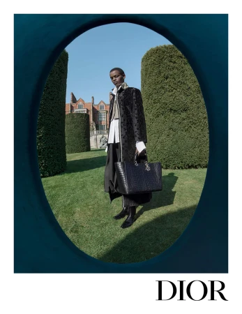 Model standing on a lawn framed by an oval archway, wearing a long dark coat and holding a black handbag.