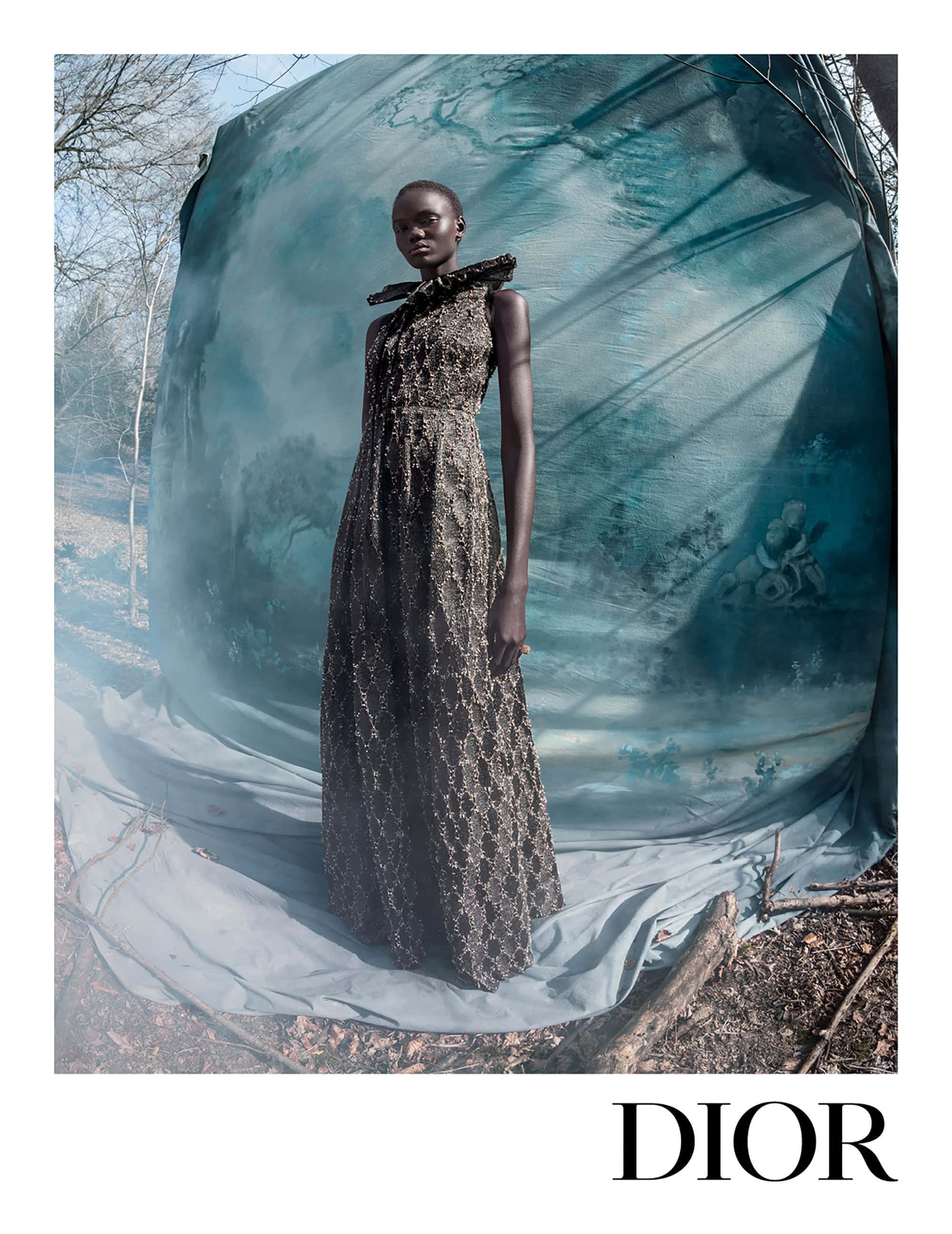Model wearing a dark textured gown standing in front of a draped teal painted backdrop outdoors.