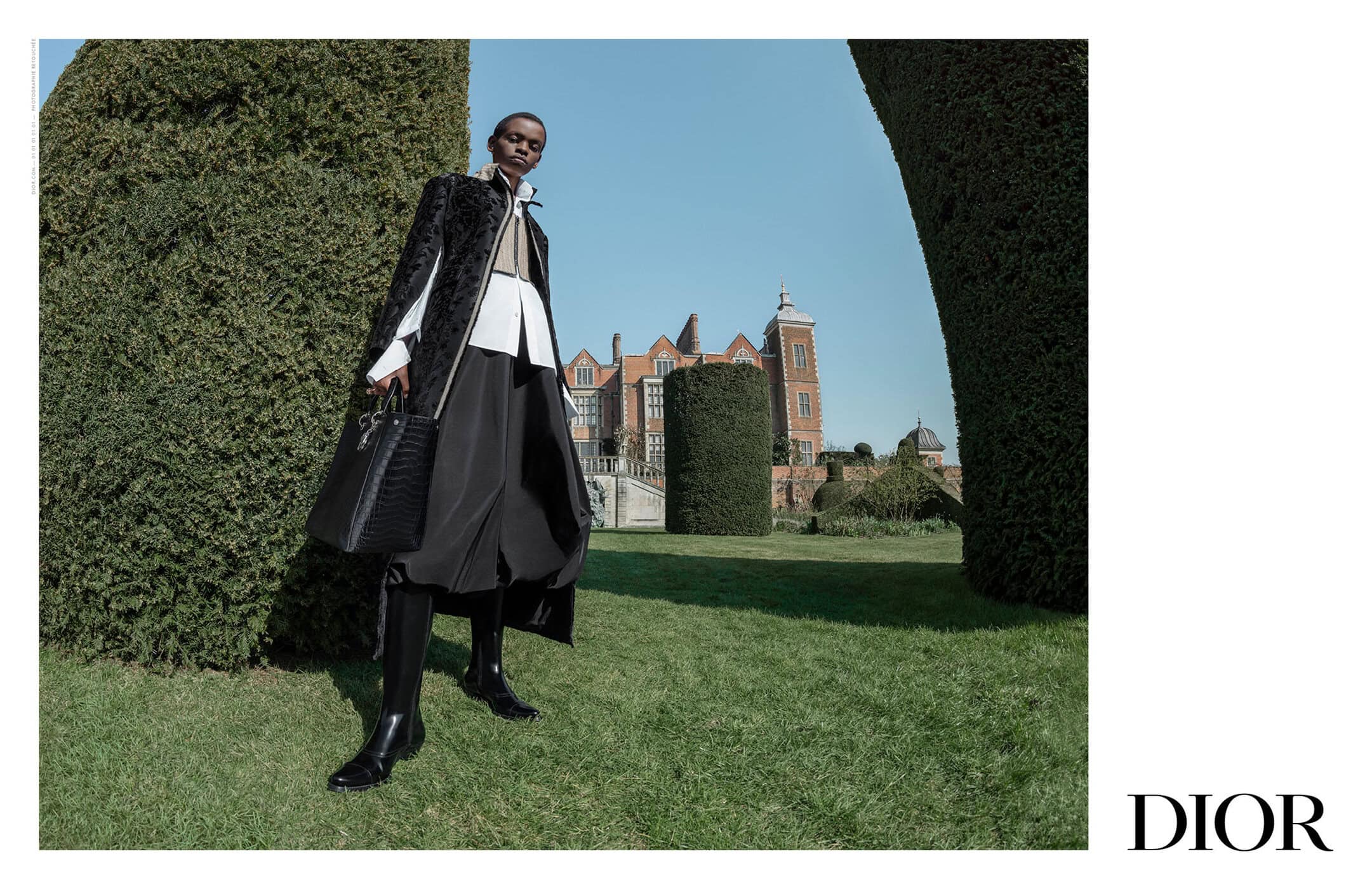 Model wearing a black coat and boots holding a handbag outdoors with topiary and a country house in the background for Dior Fall 2025 campaign.
