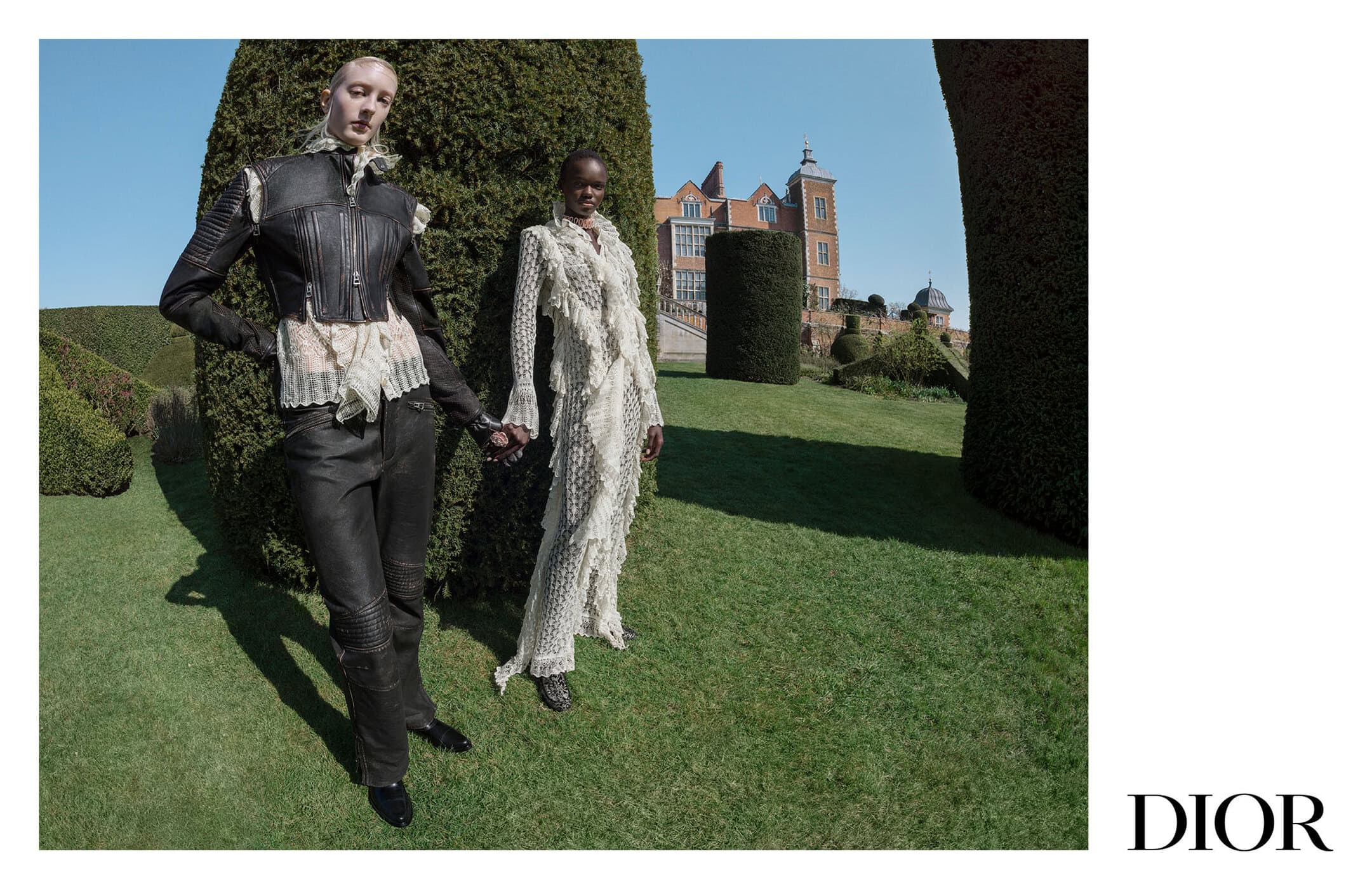 Two models standing on a lawn in front of sculpted topiary and a large brick building for a Dior advertising campaign.