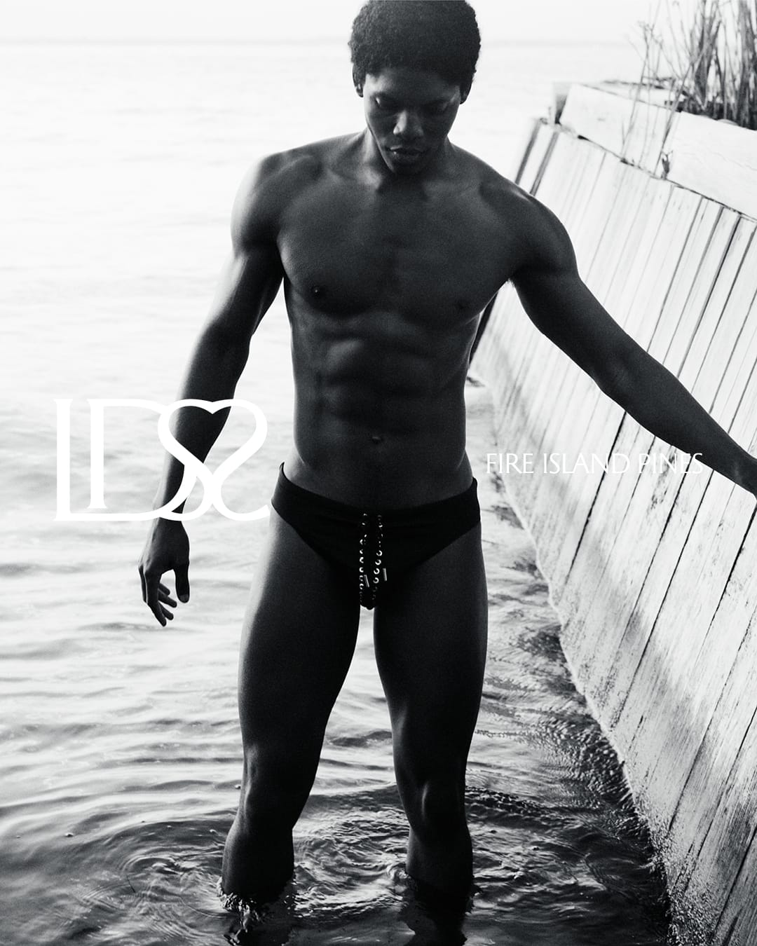 Monochrome swimwear campaign image featuring a black man standing in shallow water next to a wooden wall.