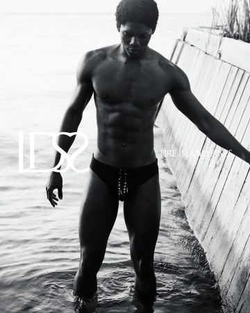 Monochrome swimwear campaign image featuring a black man standing in shallow water next to a wooden wall.