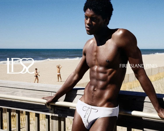 Shirtless black man wearing white lace-up swim briefs leaning on a wooden boardwalk railing at the beach