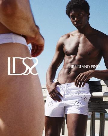 Shirtless black model wearing white swim trunks leaning on a wooden railing against a clear blue sky.