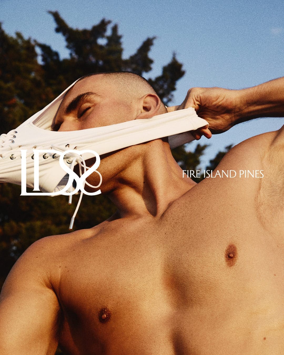 Shirtless male model wearing white lace-up briefs pulled over his face against an outdoor sky background