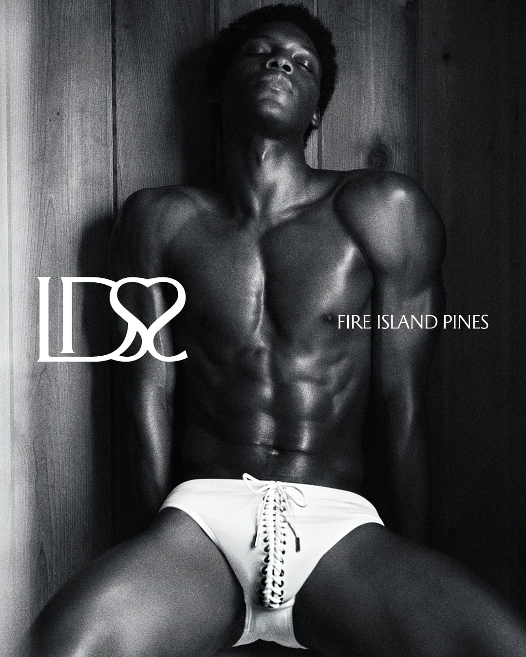 Monochrome portrait of a black man wearing white lace-up swim briefs against a wooden paneled wall