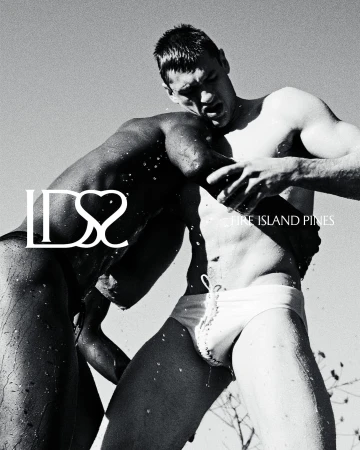 Two men wrestling outdoors in swimwear, water splashing against their skin under bright sunlight, captured in a dynamic black and white composition.