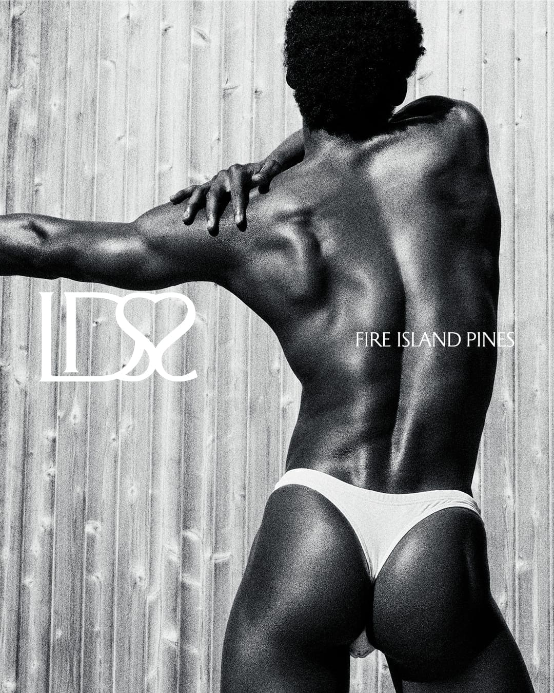 Black-and-white fashion image of a man wearing white briefs, posed with back turned against a vertical wooden slat wall.