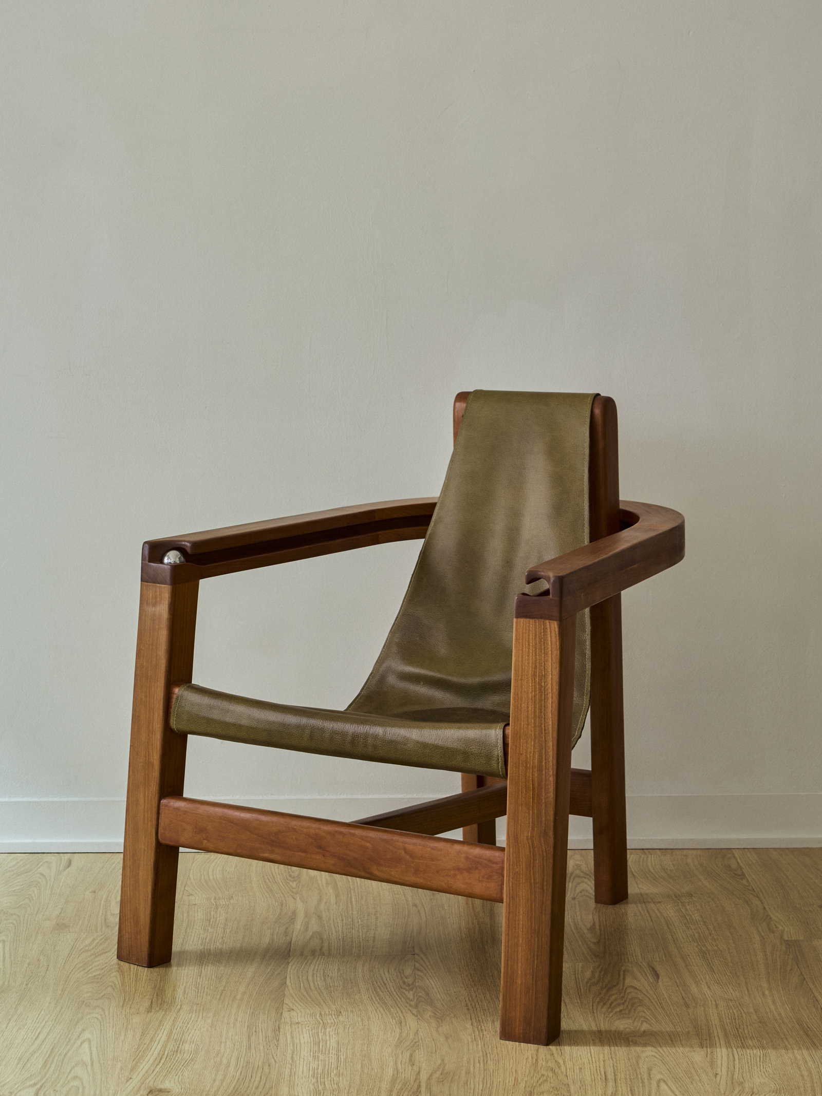 Wooden armchair with an olive green leather sling seat and backrest positioned against a pale wall.