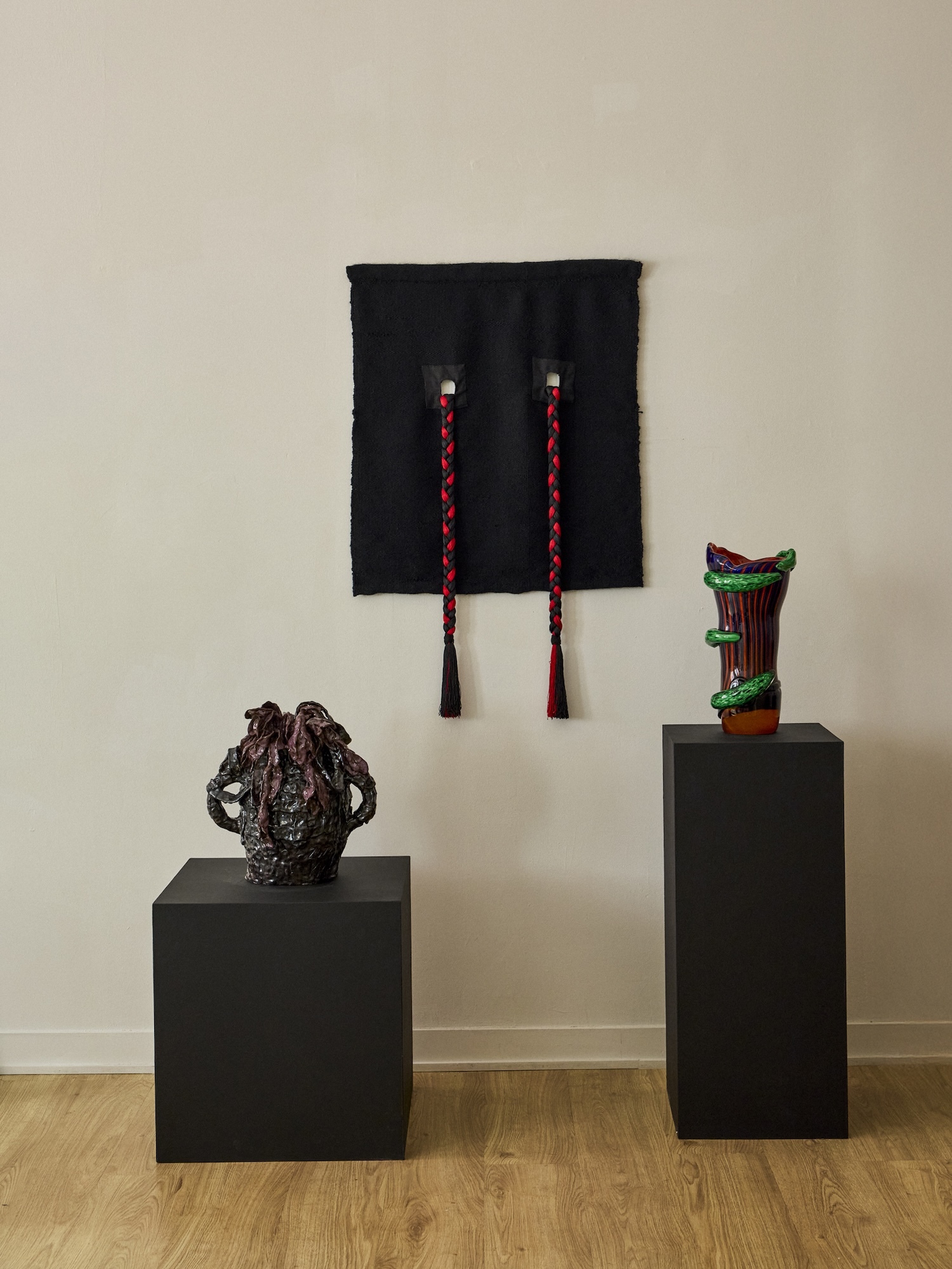 Gallery installation featuring two black pedestals holding textured dark sculptures and a black textile wall hanging with braided red cords