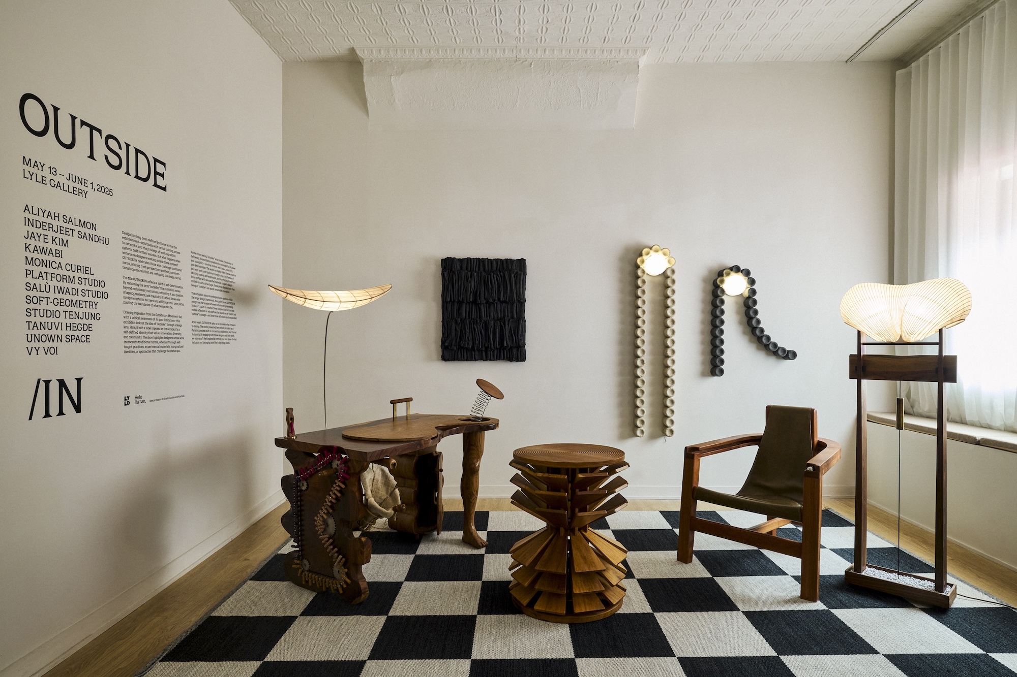 Gallery installation interior featuring modern wooden furniture, sculptural lighting, and a black and white checkered rug on the floor.