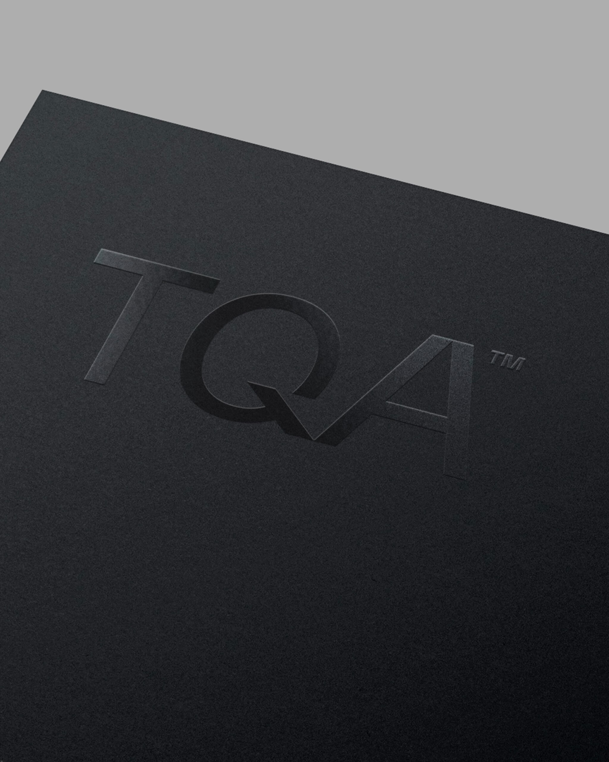 Embossed TQA brand logo on a matte black textured paper surface with minimalist lighting.