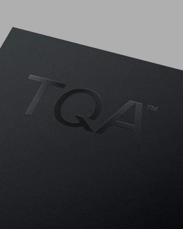 Embossed TQA brand logo on a matte black textured paper surface with minimalist lighting.