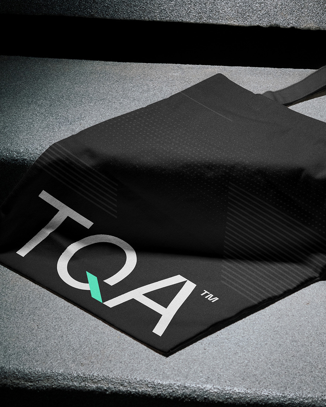 Black fabric bag with TQA logo resting on a textured concrete step surface.