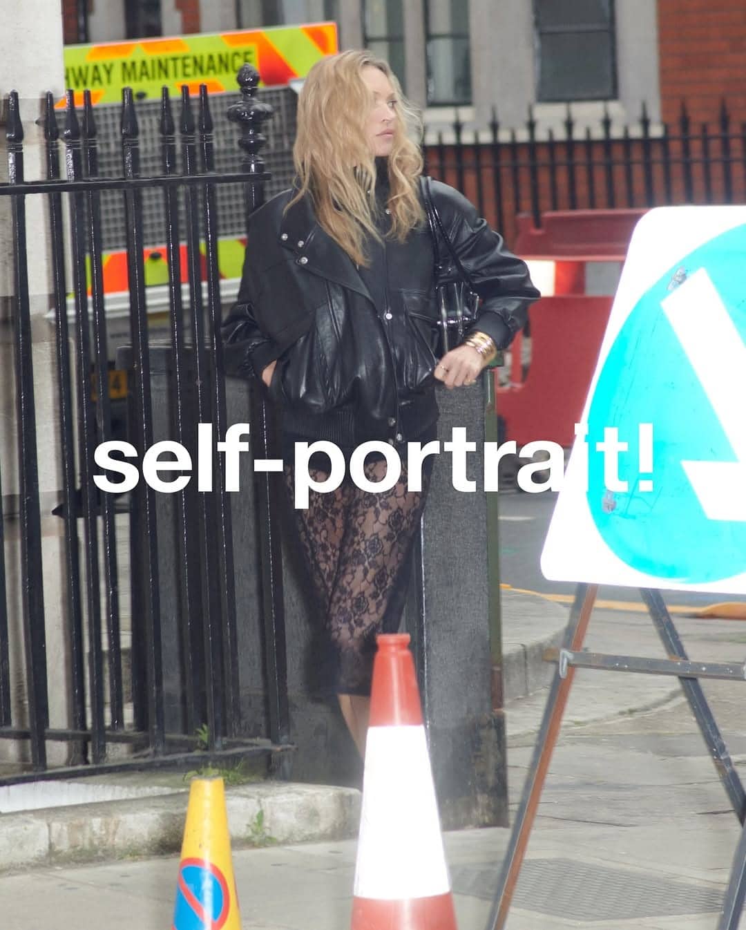 Kate Moss standing on a city street wearing a black leather jacket and sheer lace dress for Self-Portrait