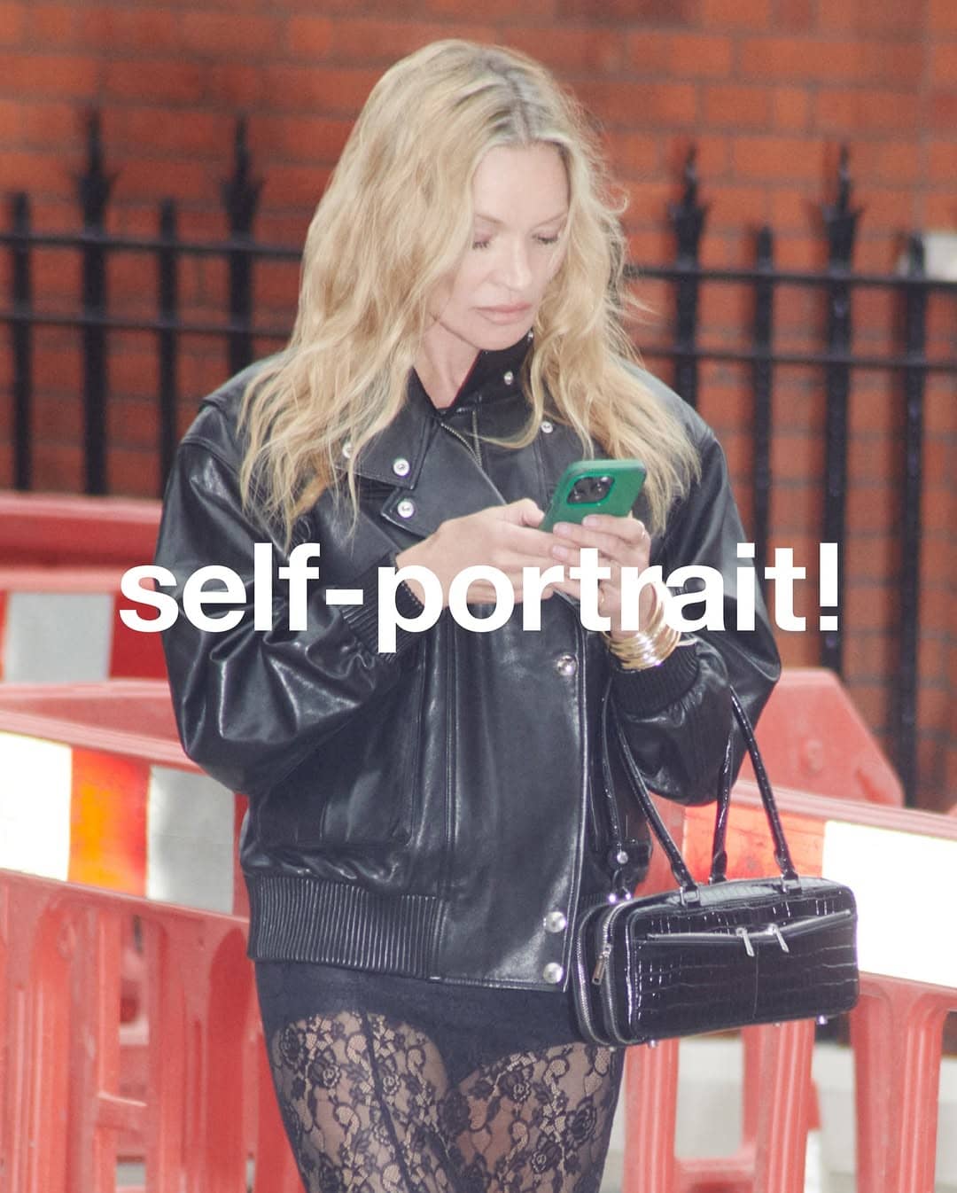 Model Kate Moss in a black leather bomber jacket holding a smartphone outdoors with a brick background