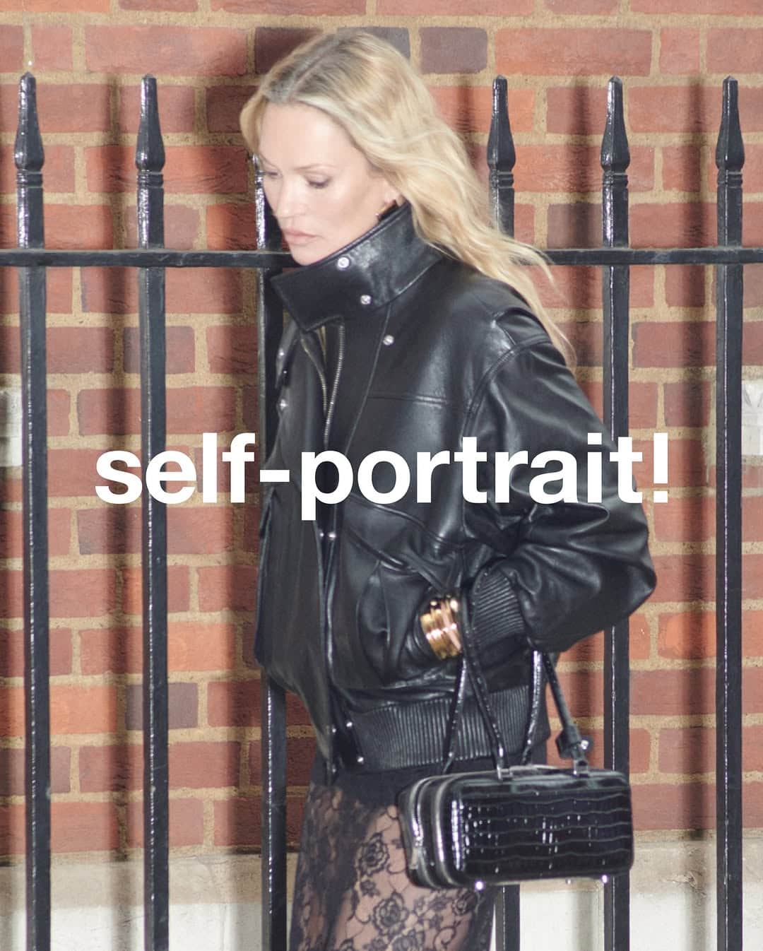 Kate Moss wearing a black leather jacket and holding a black handbag against a brick wall with iron railings.