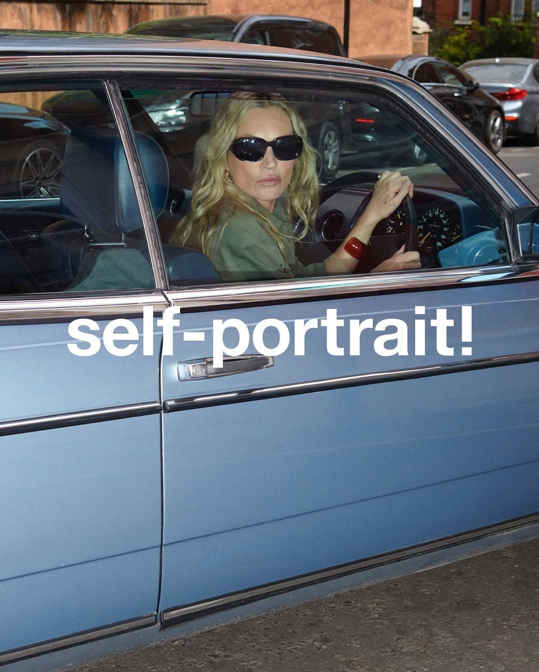 Model Kate Moss in dark sunglasses sitting behind the steering wheel of a vintage blue car for a Self-Portrait campaign.