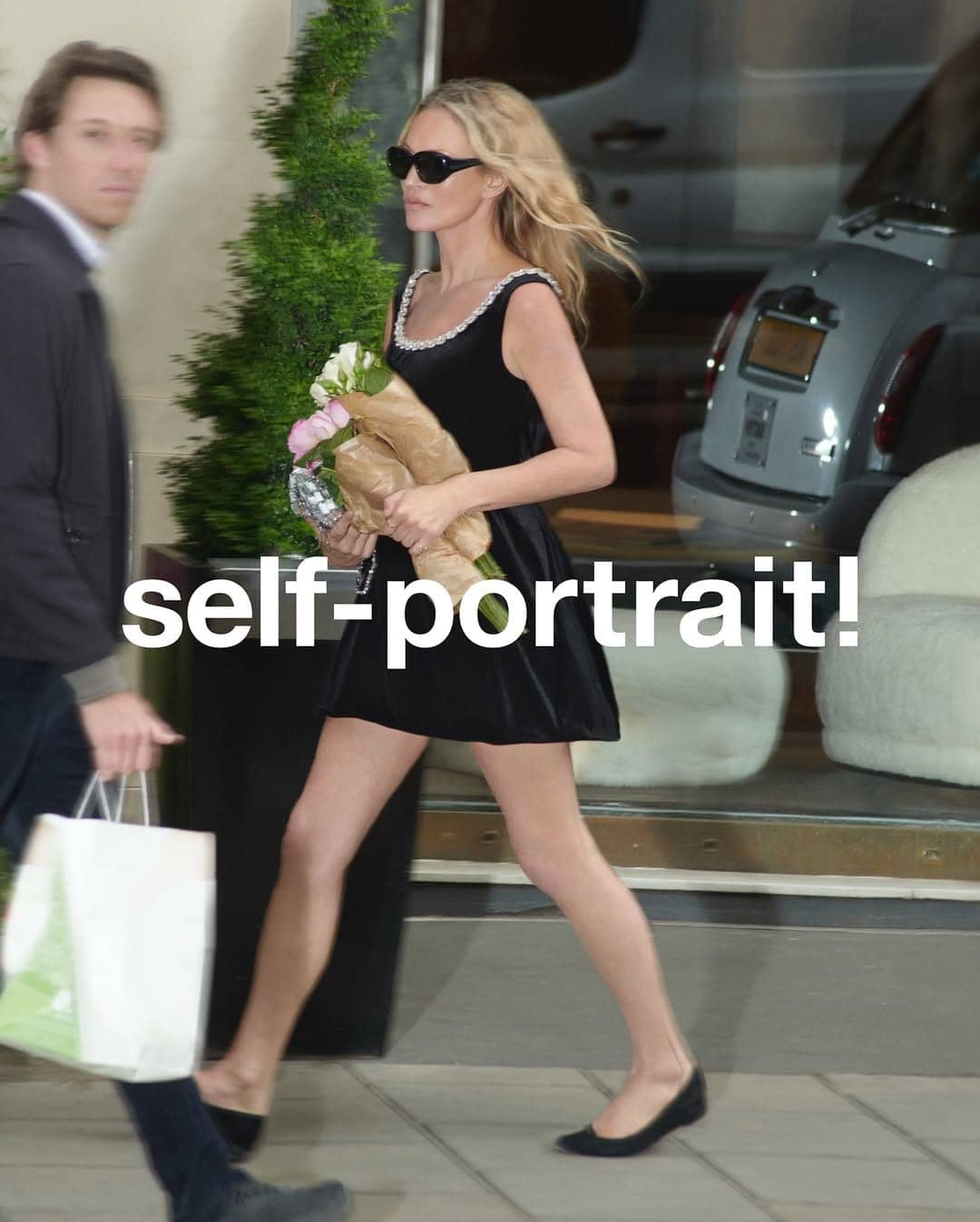 Kate Moss wearing a black dress with sunglasses holding flowers, walking past a storefront.