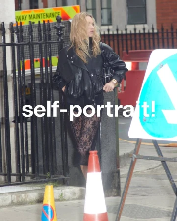 Kate Moss standing on a city street wearing a black leather jacket and sheer lace dress for Self-Portrait