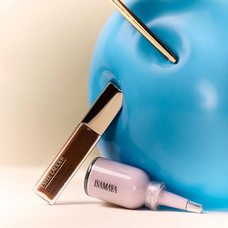 Beauty still life featuring Estee Lauder Double Wear concealer and Isamaya product with a blue apple-shaped prop on a cream backdrop.