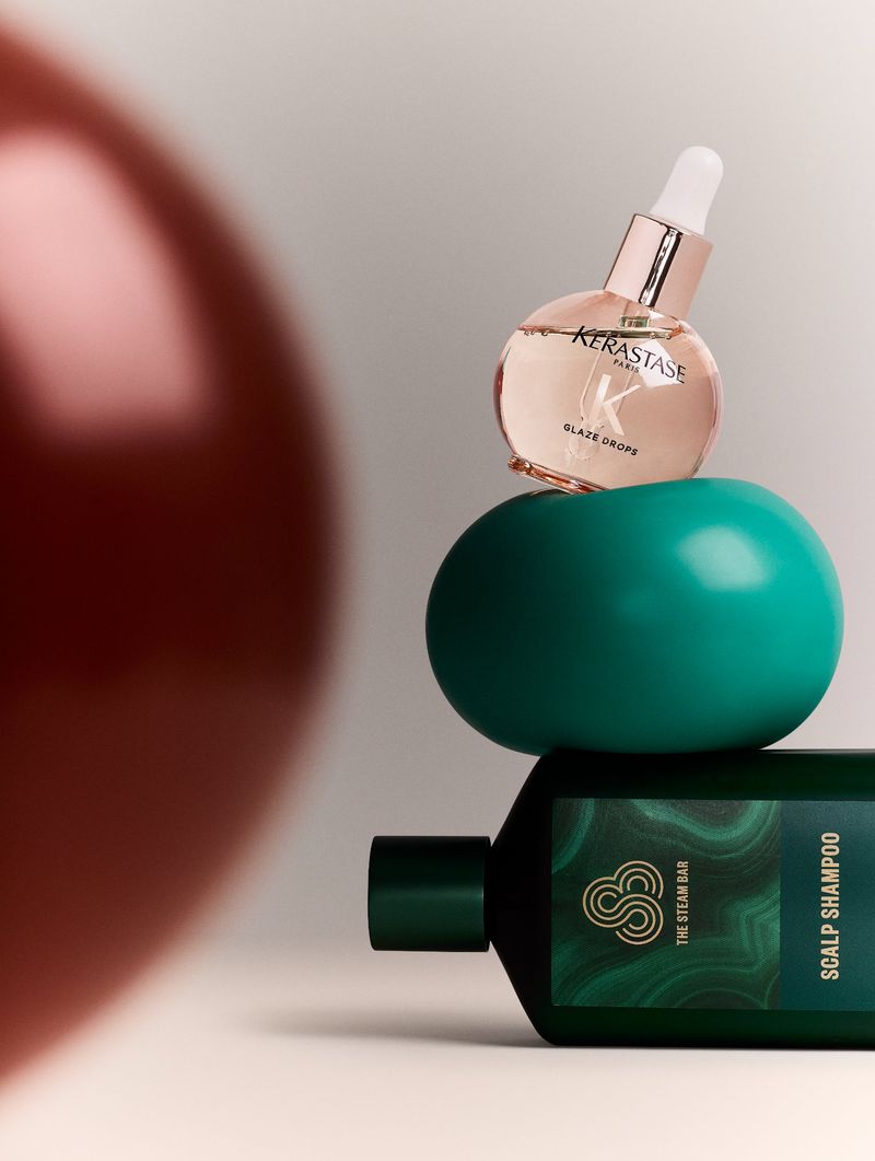 Kerastase Glaze Drops and The Steam Bar Scalp Shampoo on teal-colored sphere with a blurred reddish-brown sphere on the left.