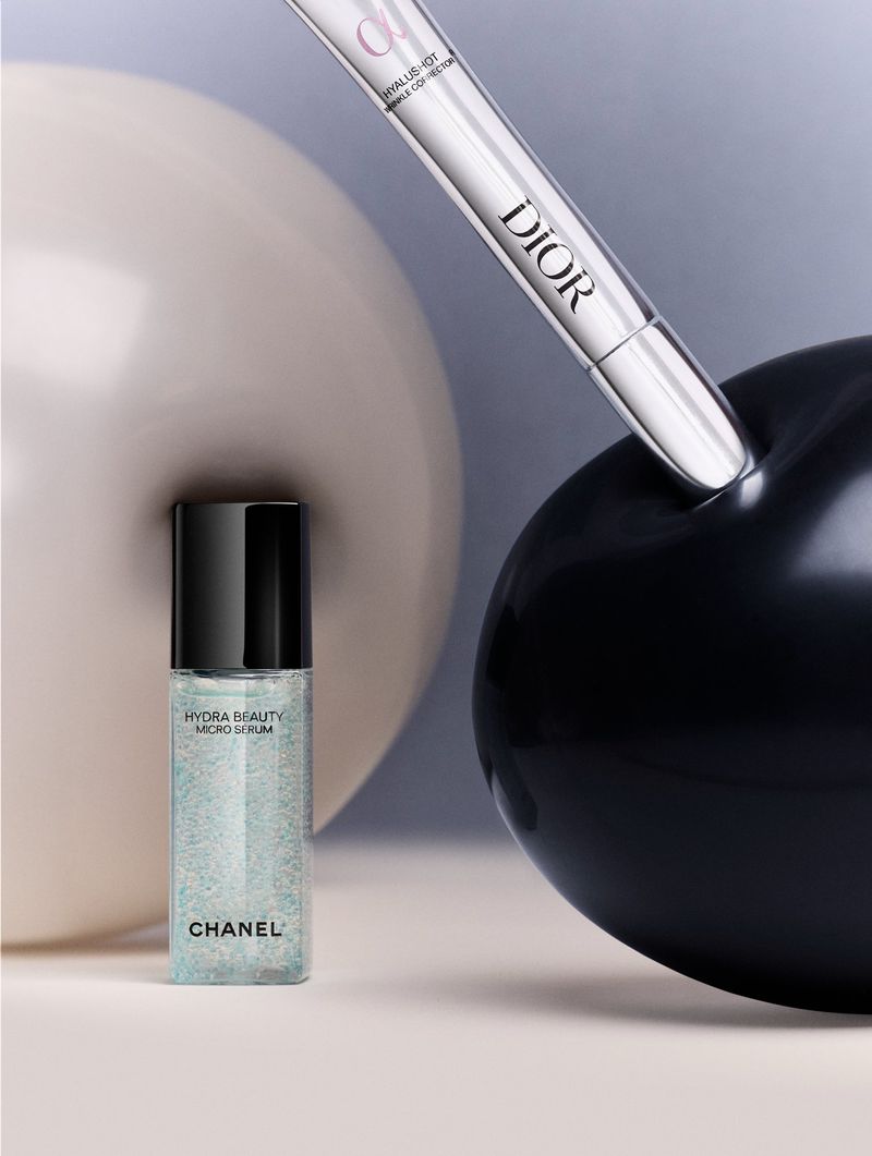 Cosmetic still life with Chanel Hydra Beauty Micro Serum and Dior concealer set against neutral and black sphere backgrounds.