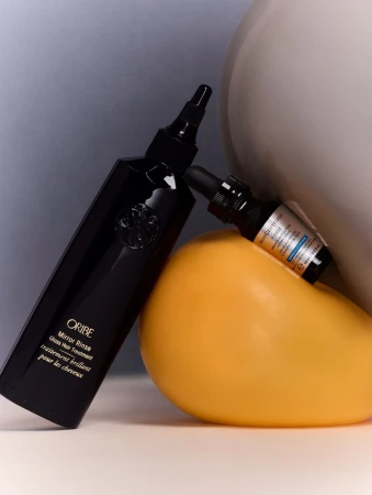Cosmetic product still life featuring Oribe hair treatment and serum bottles on a white surface with grey and yellow geometric shapes.
