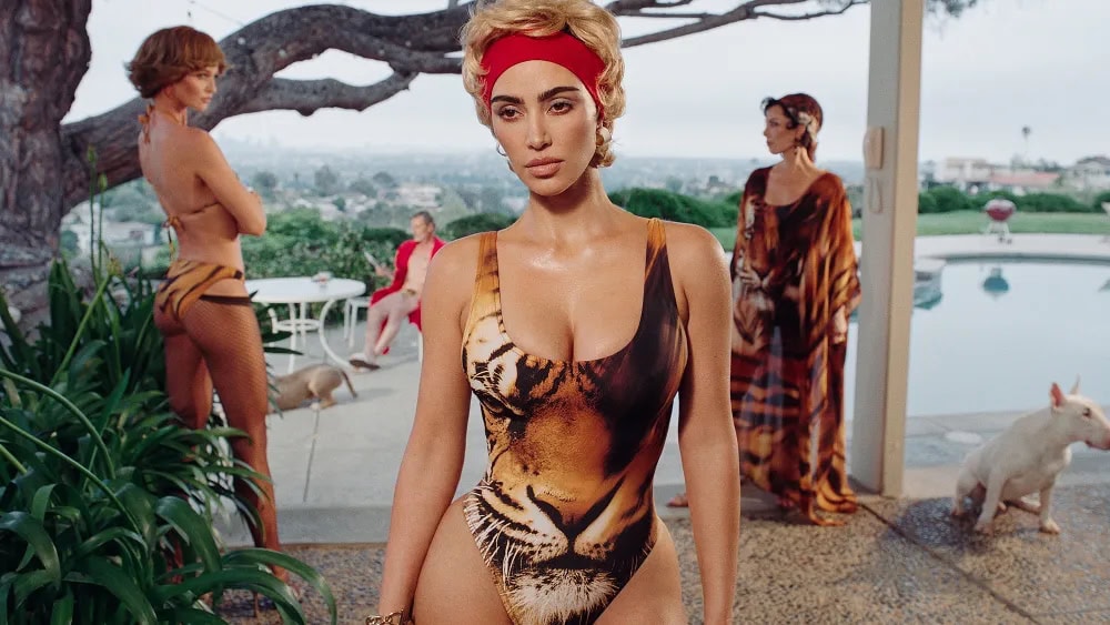 Kim Kardashian wearing a tiger print swimsuit and red headband by a swimming pool in a retro styled advertising campaign photograph