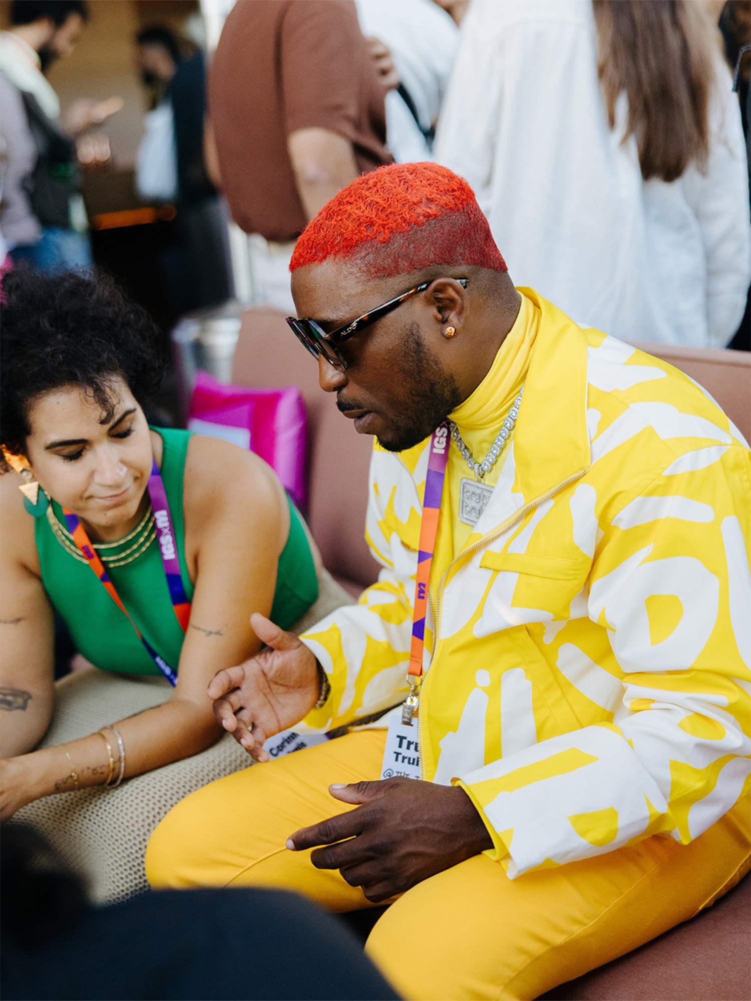 Black man with red hair wearing a yellow and white patterned jacket and yellow pants sitting and gesturing during a Manychat event