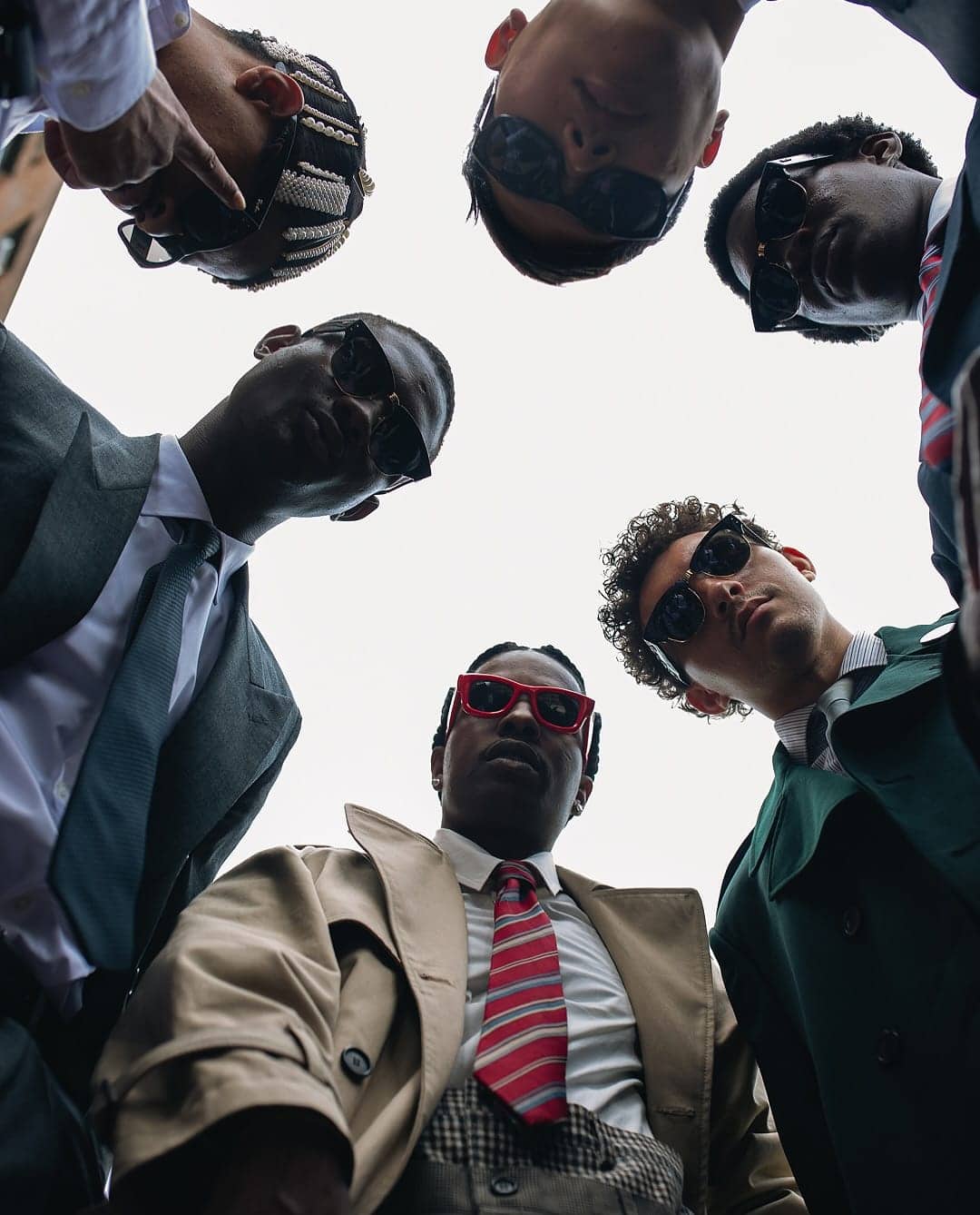 Low angle group shot of several men in suits and sunglasses looking down against a bright sky.
