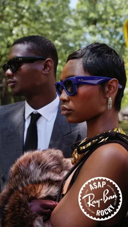 Two models wearing sunglasses outdoors, featuring a woman in purple frames and a man in black suit and sunglasses.