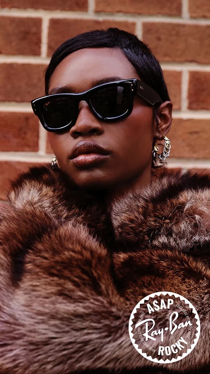 Black woman wearing dark sunglasses and a brown fur coat against a red brick wall for a Ray-Ban campaign.