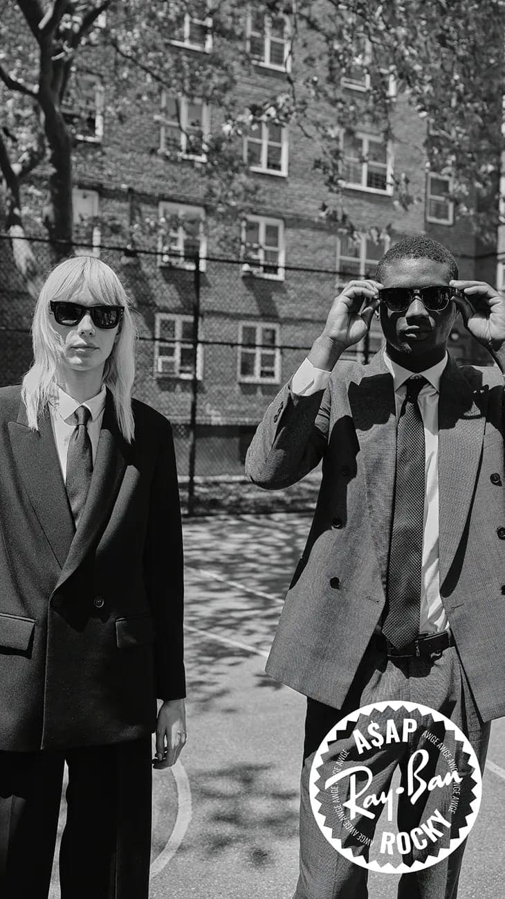 Black and white portrait of a blonde woman and a black man wearing suits and sunglasses outdoors in front of a brick building.