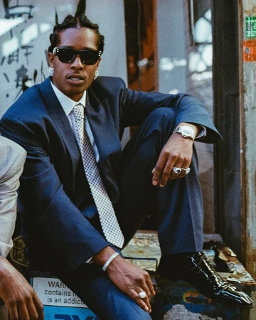 A$AP Rocky wearing a dark suit, tie, sunglasses and earrings while seated outdoors for a Ray-Ban ad campaign