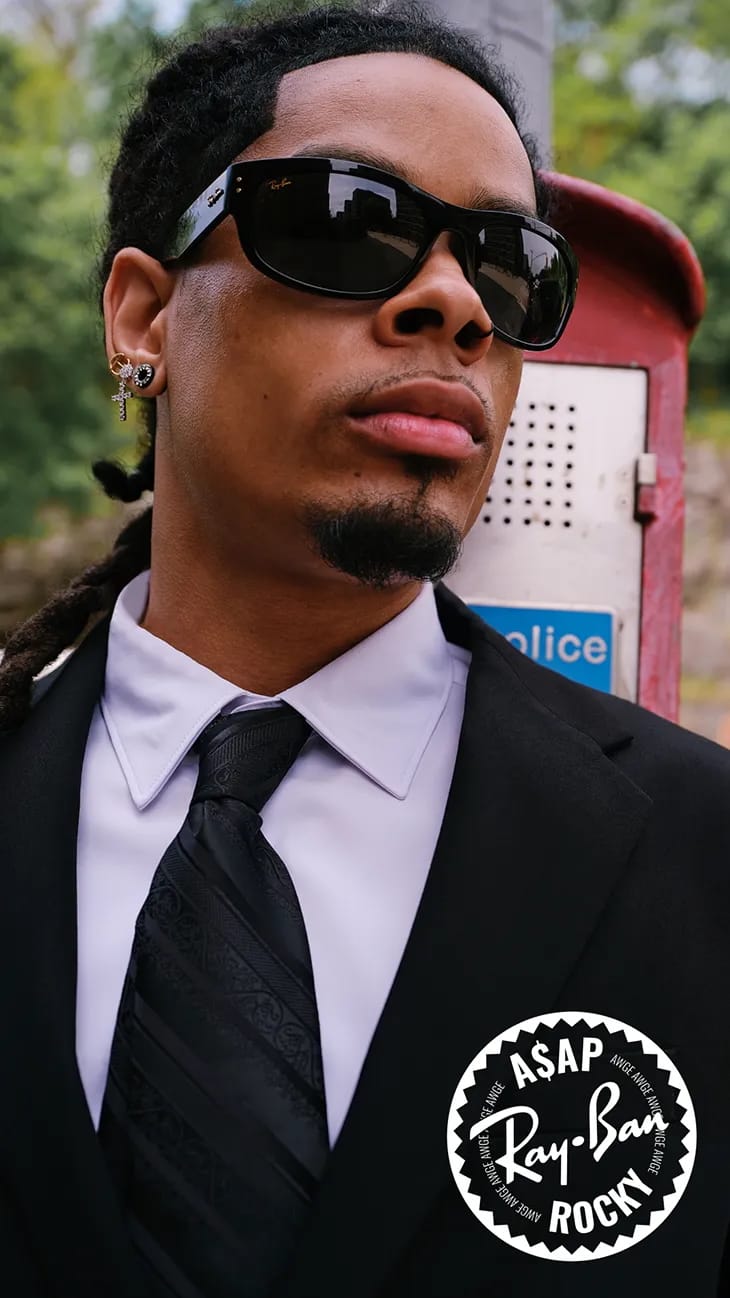 Man wearing black sunglasses, a white shirt, a patterned black tie, and a black suit jacket outdoors near a red postal box