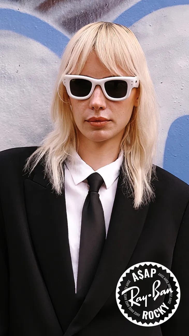 Model with blonde hair wearing white sunglasses, a white shirt, black tie, and a black suit against a colorful graffiti background.