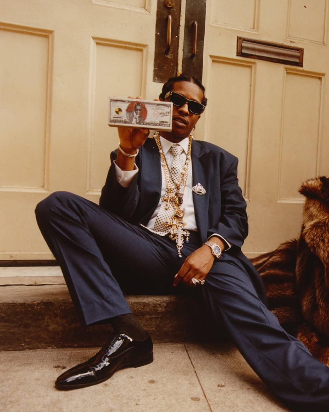 A$AP Rocky wearing a navy suit and sunglasses, seated on stone steps holding a stack of custom cash for a Ray-Ban campaign.