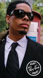 Man wearing black sunglasses, a white shirt, a patterned black tie, and a black suit jacket outdoors near a red postal box