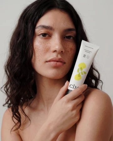 Close-up portrait of a woman with dark wavy hair and freckles holding a CÎME skincare tube against a neutral background.