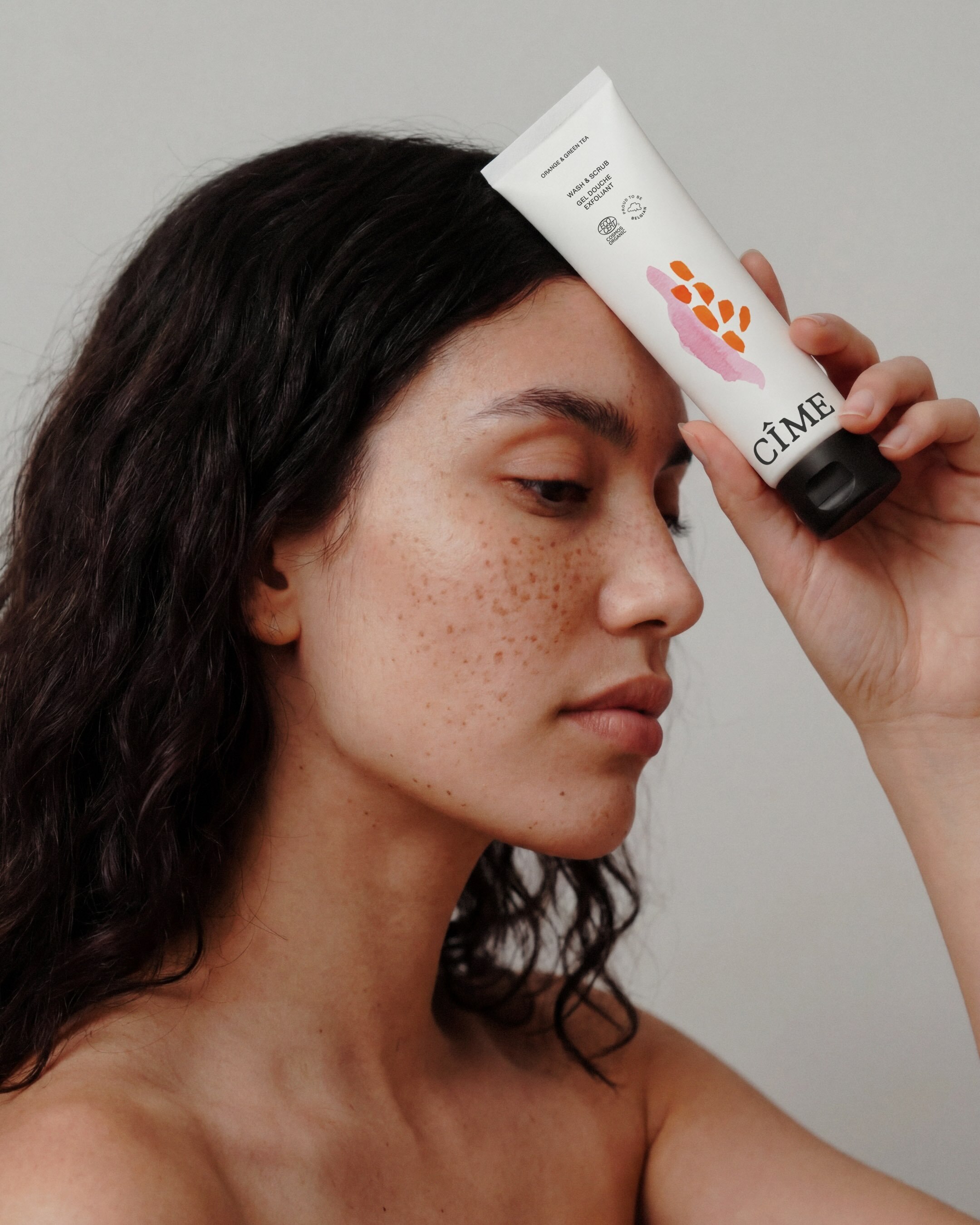 Close-up portrait of a woman with freckles holding a white CÎME skincare tube against her forehead against a plain grey background