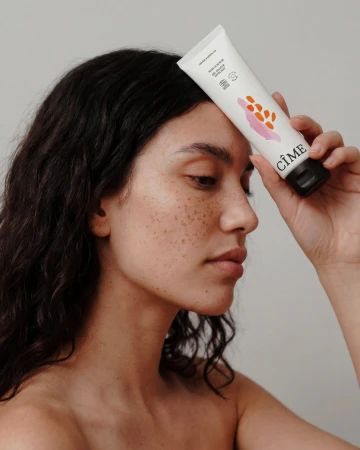 Close-up portrait of a woman with freckles holding a white CÎME skincare tube against her forehead against a plain grey background