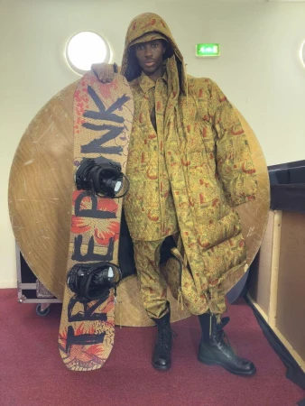 Model wearing patterned hooded coat and matching trousers holding a branded snowboard for Vivienne Westwood campaign