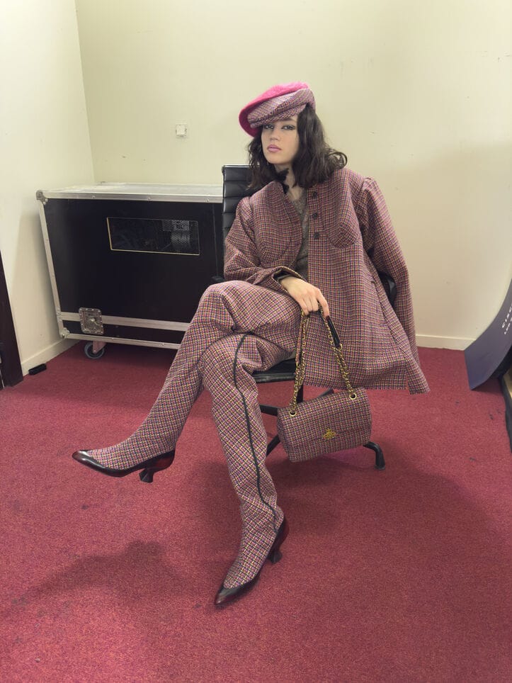 Model seated in an office chair wearing a pink plaid suit, matching hat, boots, and handbag against a neutral wall.