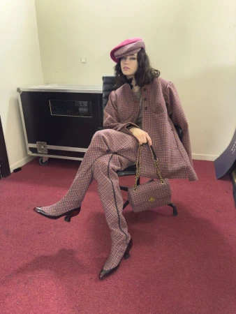 Model seated in an office chair wearing a pink plaid suit, matching hat, boots, and handbag against a neutral wall.