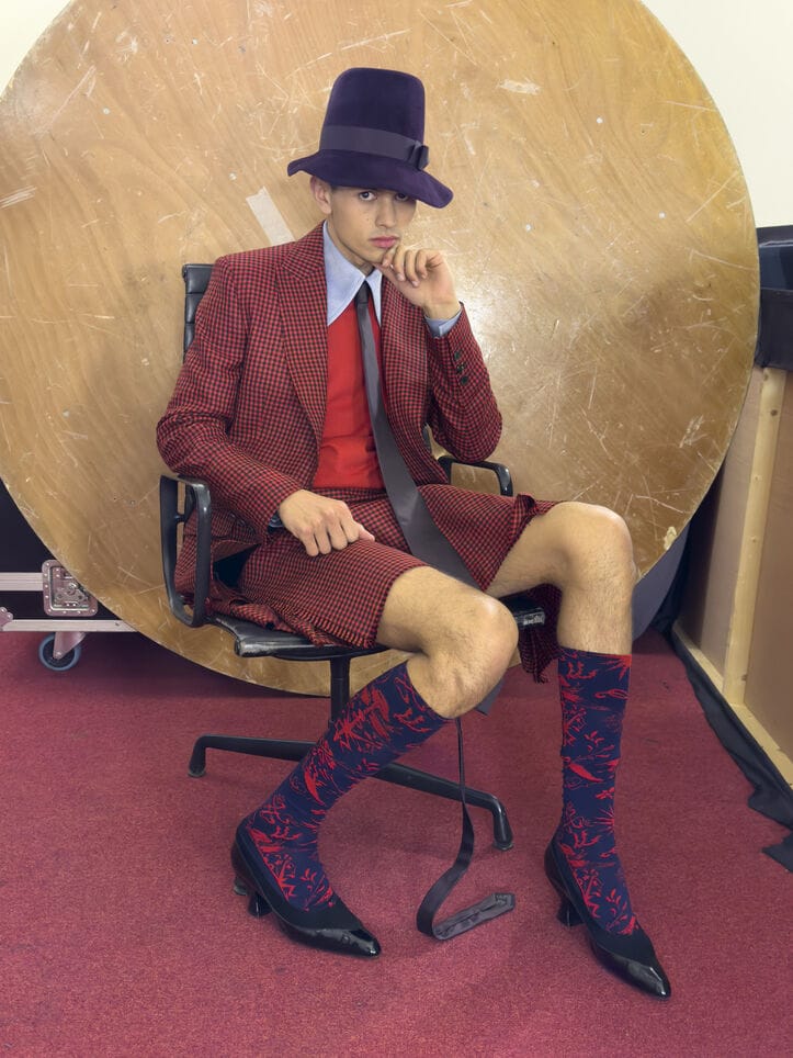 Model wearing a patterned red suit, purple top hat, and patterned blue socks seated on a rolling chair