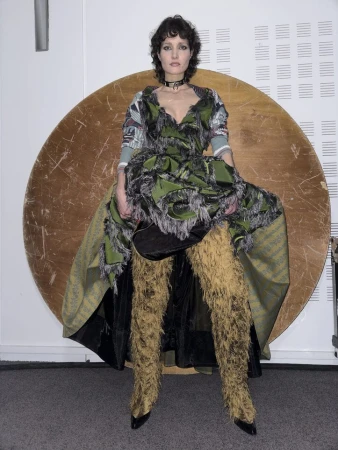 Model wearing a textured green dress and tall furry boots standing in front of a large circular wooden disc.