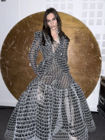 Model wearing a patterned sheer gown posing in front of a large golden circular disc for Vivienne Westwood