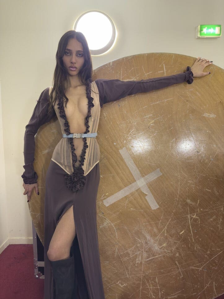 Model leaning against a large wooden disc wearing a plunging dress with ruffles and dark boots for Vivienne Westwood.